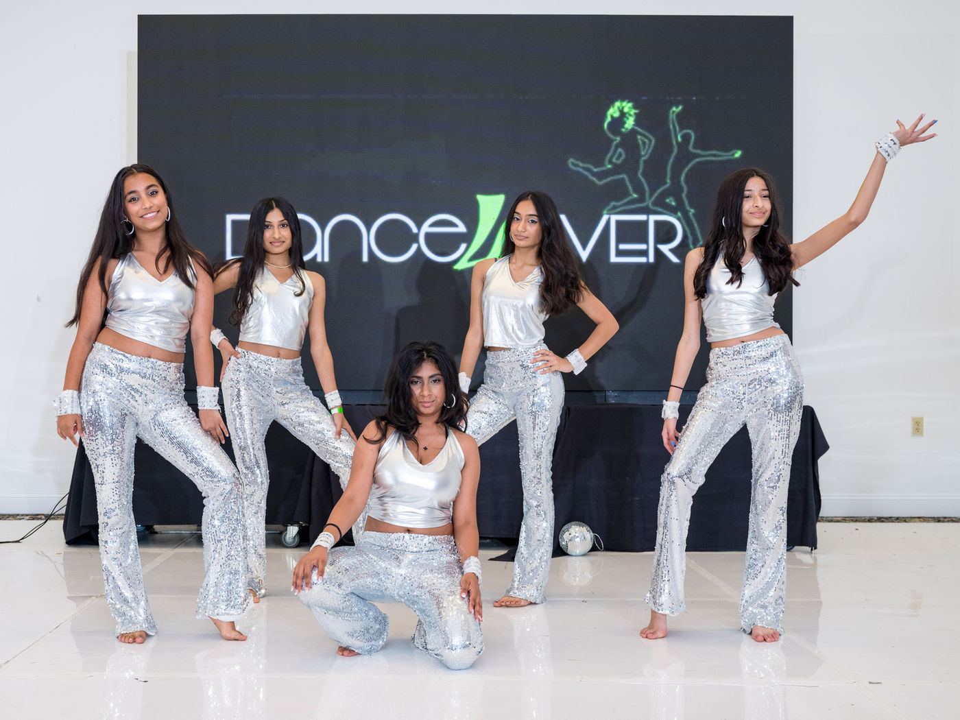 Five Dance4Ever dancers in silver costumes posing in front of the studio banner